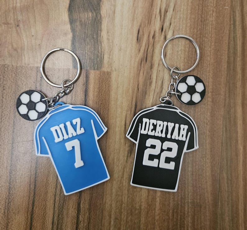 May include: Two soccer jersey keychains with a soccer ball charm. The blue jersey has the name "DIAZ" and the number "7" on the back. The black jersey has the name "DERIYAH" and the number "22" on the back.
