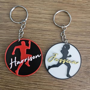 May include: Two personalized keychains. One features a red runner silhouette on a black circle with the name "Harrison" in white script. The other has a black runner silhouette on a white circle with the name "Jessica" in gold script.