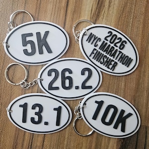 May include: Five oval white keychains with black borders and raised black lettering. The keychains feature the text 5K, 26.2, 13.1, 10K, and 2026 NYC MARATHON FINISHER. Each keychain includes a silver metal split ring attached by a short chain.