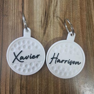 May include: Two white golf ball-shaped keychains with black text. One keychain says "Xavier" and the other says "Harrison". Each keychain has a silver metal ring attached for keys.