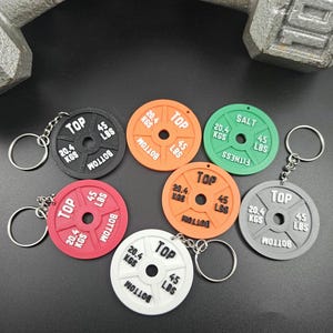 May include: Six different weight plate keychains, each with a black and white design. The keychains are made of plastic and feature the words "TOP", "BOTTOM", "KGS", and "LBS".