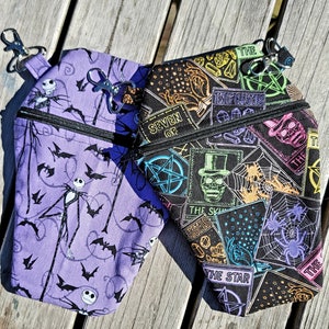 May include: Two purple and black zippered pouches with different designs. The first pouch has a purple background with a white outline of a skeleton figure and bats. The second pouch has a black background with colorful tarot card designs and a pentagram.