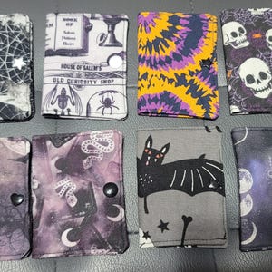 May include: Eight fabric wallets with various spooky designs. The wallets feature patterns like spiderwebs, skulls, bats, and celestial motifs. Each wallet has a different color scheme, including black, purple, and orange.