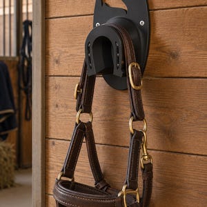 Op de afbeelding: Een zwarte wandhouder met een uitgesneden silhouet van een paardenkop en een hoefijzervormige haak. Een donkerbruin lederen paardenhalster met messingkleurige metalen gespen, ringen en beslag hangt stevig aan de haak aan de muur.