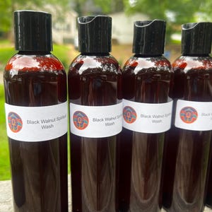 May include: Several dark brown bottles with black caps are arranged in a row. Each bottle has a white label with the text "Black Walnut Spiritual Wash" and a circular design. The bottles are filled with a dark liquid.