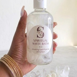 Puede incluir: Un frasco de plástico transparente con tapa blanca que contiene un líquido y elementos florales blancos. La etiqueta circular presenta una letra S grande decorada con flores, el texto SPIRITUAL WHITE BATH, 8 oz, y cleansing/purification and peace.