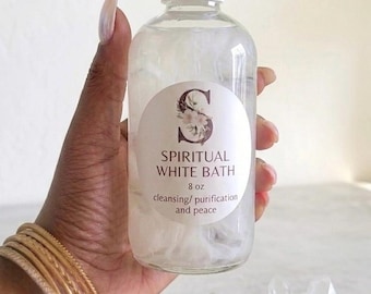 May include: A clear plastic bottle with a white cap containing a liquid and white floral elements. The circular label features a large letter S decorated with flowers, the text SPIRITUAL WHITE BATH, 8 oz, and cleansing/purification and peace.