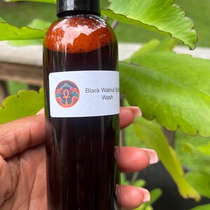 May include: A clear bottle with a black cap, filled with a dark brown liquid. The label reads "Black Walnut Soap Wash" with a colorful graphic. The bottle is held in front of green foliage.