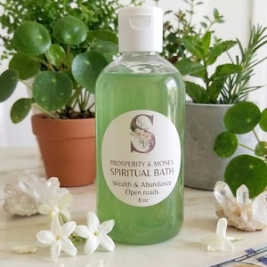 May include: A clear plastic bottle containing green liquid with a white cap and a label reading Prosperity & Money Spiritual Bath Wealth & Abundance Open roads 8 oz. The bottle is positioned with white flowers and a 100 dollar bill.
