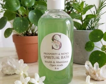 May include: A clear plastic bottle containing green liquid with a white cap and a label reading Prosperity & Money Spiritual Bath Wealth & Abundance Open roads 8 oz. The bottle is positioned with white flowers and a 100 dollar bill.