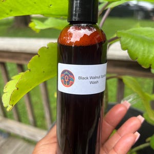 May include: A clear bottle filled with a dark liquid, labeled "Black Walnut Spirit Wash." The bottle has a black cap and a white label with a colorful design. The bottle is held in a hand, with green foliage in the background.