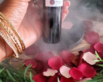 May include: A black glass bottle with a white label is held in front of a background of red and white rose petals and green foliage. The bottle is surrounded by a mist. The label reads "Black Tourmaline Oil".