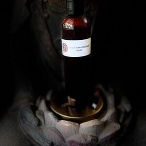 May include: A brown bottle with a black cap, labeled "Black Walnut Spiritual Wash." The bottle is set against a dark background, resting on a golden-colored surface, and is likely a skincare or wellness product.