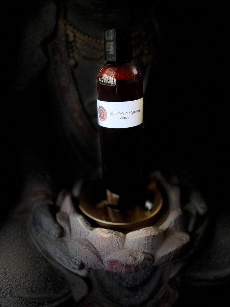 May include: A brown bottle with a black cap, labeled "Black Walnut Spiritual Wash." The bottle is set against a dark background, resting on a golden-colored surface, and is likely a skincare or wellness product.