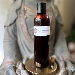 May include: A dark brown bottle with a black cap and a white label that reads "Black Walnut Spiritual Wash." The bottle is held in front of a statue of a Buddha. The liquid inside the bottle is a deep reddish-brown color.