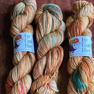 May include: Three skeins of yarn in shades of brown, cream, and green with a speckled effect. Each skein has a label that reads "Eye Candy Fiber" and "Hand dyed in the Pacific Northwest".