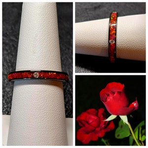 May include: A black ring with a vibrant orange inlay and a small, clear gemstone. The ring is displayed on a white surface, showcasing its design. A red rose is also visible in the image.