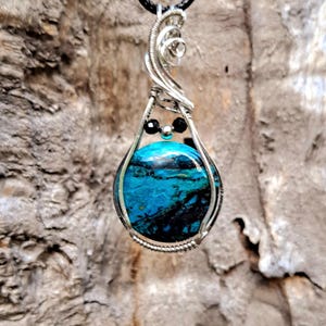 May include: A handcrafted pendant featuring a vibrant blue and black gemstone encased in a silver wire wrap. The pendant hangs from a black cord. The gemstone has a unique pattern, resembling an abstract landscape.