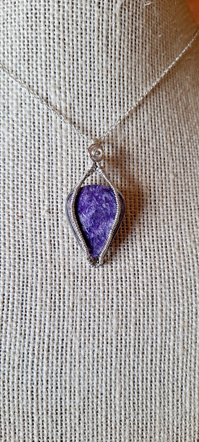 May include: A silver wire wrapped pendant featuring a purple and white gemstone. The pendant is hanging from a silver chain on a fabric background.