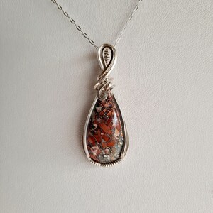 May include: A silver wire wrapped pendant featuring a teardrop-shaped red, brown, and gray stone.