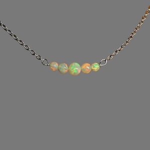 May include: A delicate necklace featuring a thin silver-toned metal chain with five round, iridescent opal beads arranged in a horizontal row at the center. The opals display flashes of green, orange, and yellow light against a milky white base.