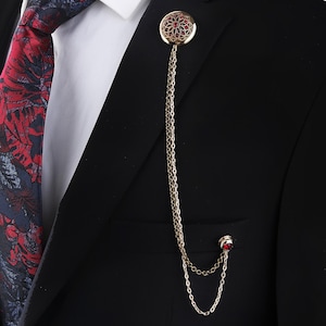 May include: A black suit jacket with a red and blue patterned tie and a gold chain with a red jeweled pin.