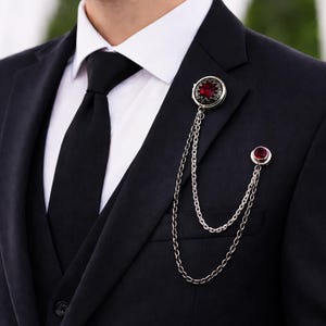 Może przedstawiać: Srebrna przypinka do klapy z okrągłym elementem centralnym z ciemnoczerwonym kamieniem i misternymi detalami metalowymi, połączona dwoma drapowanymi srebrnymi łańcuszkami z mniejszą, pasującą przypinką z pojedynczym czerwonym kamieniem.