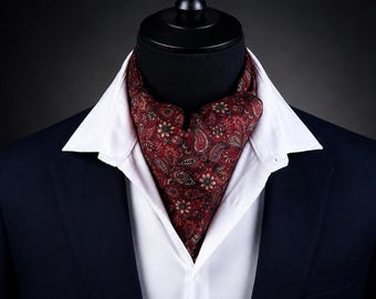 Könnte beinhalten: Ein dunkelrotes Ascot-Tuch mit einem detaillierten Paisley- und Blumenmuster in Schwarz-, Weiß- und Grautönen. Das Ascot wird in ein weißes Hemd mit Kragen gesteckt, das unter einem dunkelblauen Blazer getragen wird.