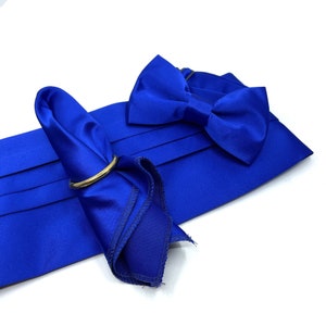 May include: A royal blue satin cummerbund with horizontal pleats, a matching pre-tied bow tie, and a folded pocket square secured by a circular gold-toned metal ring. The set features a smooth, reflective fabric finish throughout all components.