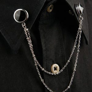 以下が含まれることがあります： 二重のチェーンで繋がれた2つの円形の黒い石のアクセントが特徴のシルバーカラーのカラーピン。このアクセサリーは黒い襟付きシャツの襟先に取り付けられており、シャツには明るい色のボタンが1つ見えています。