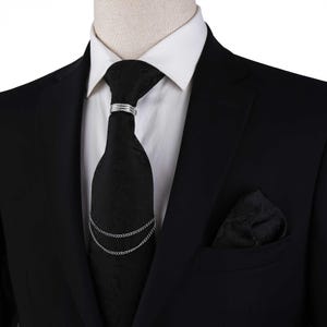 May include: A black tie with a paisley pattern, secured with a silver tie clip and chain, worn with a black suit and white dress shirt. A matching black pocket square is in the suit pocket. Formal wear.