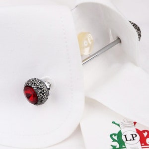 Op de afbeelding: Een paar manchetknopen met een zilveren basis en een rood kristallen middenstuk. De manchetknopen zijn omgeven door kleine zwarte stenen.