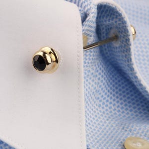 May include: A gold-toned metal collar bar featuring a black circular stone at each end, secured through the white collar of a light blue and white micro-check patterned shirt. The accessory connects the two sides of the shirt collar together.