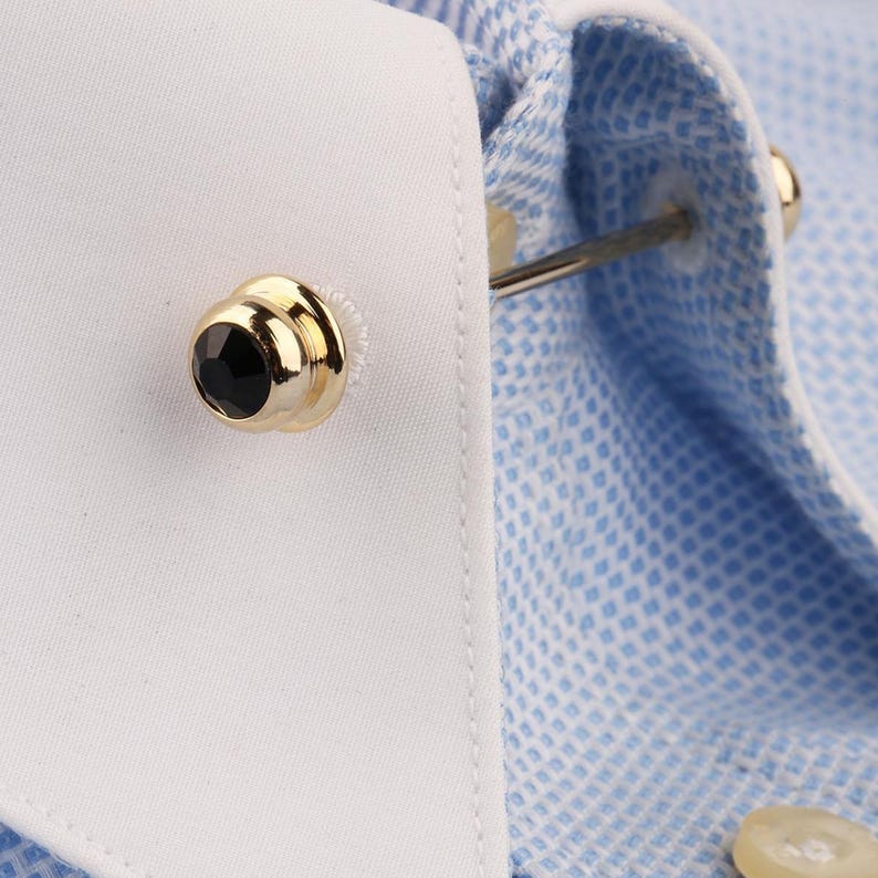 May include: A gold-toned metal collar bar featuring a black circular stone at each end, secured through the white collar of a light blue and white micro-check patterned shirt. The accessory connects the two sides of the shirt collar together.