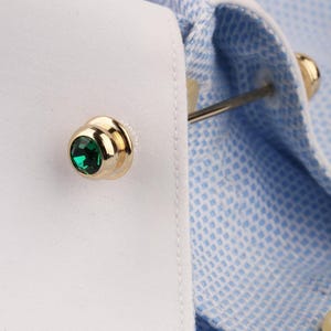 May include: A gold-toned metal cufflink featuring a round, faceted emerald green stone set in the center. The cufflink is attached to a white shirt cuff, which is paired with a light blue and white micro-patterned fabric sleeve detail.