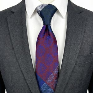 May include: A patterned necktie with a color scheme of deep red, blue, and gray. The tie features a floral design and is worn with a white collared shirt and a gray suit jacket. The tie's design is visible from the knot down.
