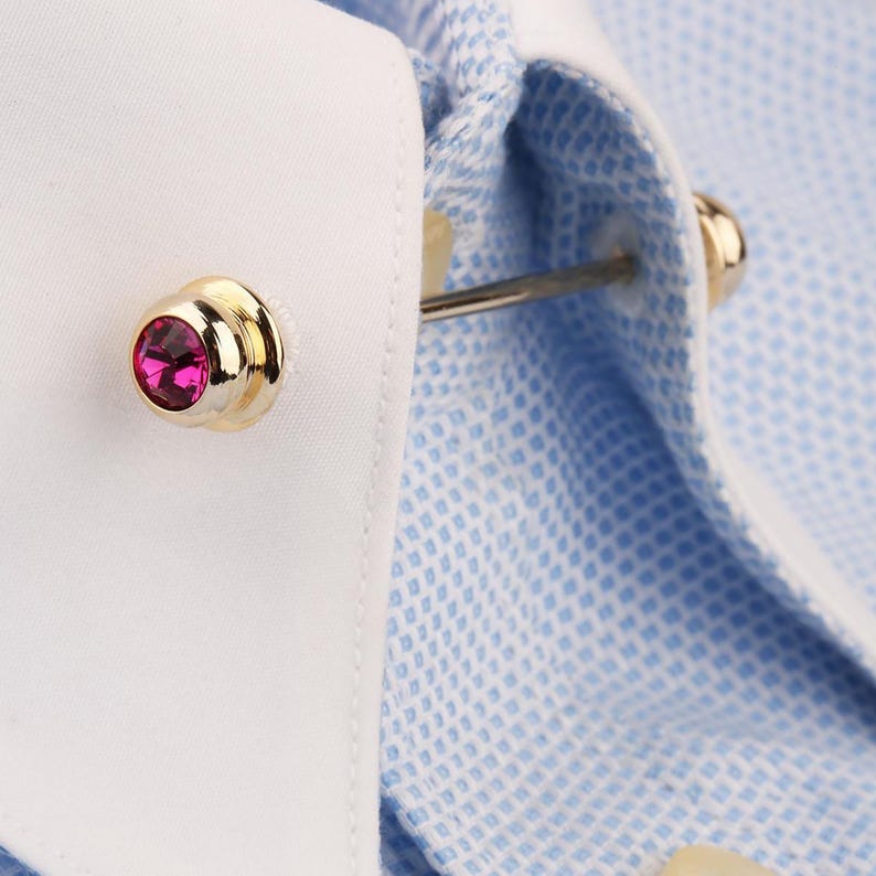May include: A gold-toned collar bar featuring a round, faceted fuchsia gemstone on each end, secured through the white collar of a light blue and white micro-check patterned dress shirt. The metal bar connects the two sides of the shirt collar.