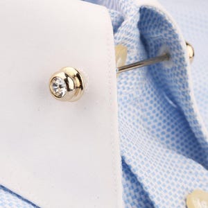 May include: A gold-toned metal collar bar featuring a clear, round crystal at each end, secured through the white collar of a light blue and white micro-check patterned dress shirt. The accessory connects the two sides of the shirt collar.