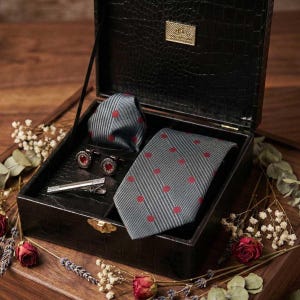 May include: A black leatherette box set containing a gray tie with red polka dots, a matching pocket square, red gemstone cufflinks, and a silver tie clip. The set is displayed on a wooden surface with floral accents.