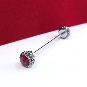 May include: A straight barbell body jewellery piece featuring a silver-toned metal shaft and two spherical ends. Each end is encrusted with small, shimmering clear crystals and features a prominent, faceted, circular red gemstone at the centre.