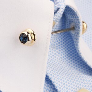 May include: A gold-toned collar bar featuring a round, faceted dark blue crystal at each end, secured through the points of a white-collared, light blue and white micro-check patterned dress shirt to hold the collar points together.