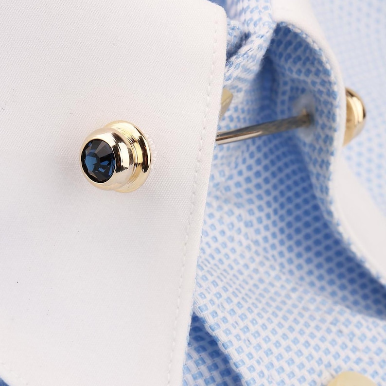 May include: A gold-toned collar bar featuring a round, faceted dark blue crystal at each end, secured through the points of a white-collared, light blue and white micro-check patterned dress shirt to hold the collar points together.