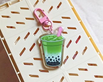 May include: An acrylic keychain featuring a green matcha bubble tea drink with dark boba pearls at the bottom, a pink straw, and a pink lobster clasp. The illustration shows a gradient green liquid with ice cubes inside a clear plastic cup.