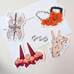 May include: Set of five stickers featuring various illustrations. One sticker shows a pair of angel wings with a sword wrapped in barbed wire. Another sticker shows a hand with vines growing around it. A third sticker shows a wreath of orange flowers. A fourth sticker shows a pair of red horns with orange flowers. The final sticker shows the text "jackfruit sketches" in a handwritten font.