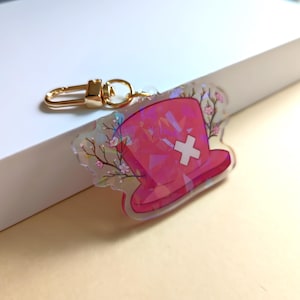 May include: A pink hat-shaped keychain with a white cross and floral accents. The keychain has a gold-colored clasp. The hat design has a geometric pattern. The overall design is translucent.