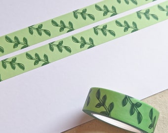 May include: A roll of light green washi tape featuring a repeating dark green vine and leaf pattern, shown alongside two strips of the same tape applied to a white surface, displaying the continuous botanical design clearly.