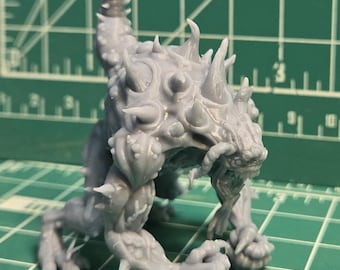 May include: A gray, unpainted resin miniature of a quadrupedal creature with a hunched posture, sharp spikes protruding from its back and limbs, a long tail, and a snarling face with visible fangs, standing on a green cutting mat with grid lines.