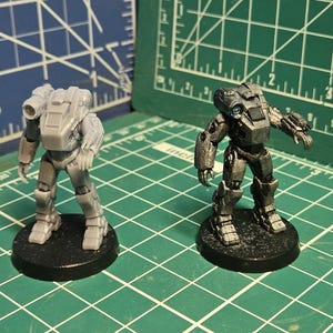 May include: Two miniature robot figurines standing on circular black bases. One figure is unpainted light gray plastic, while the other is painted in metallic silver and dark gray tones. Both robots feature articulated limbs and mounted weaponry.