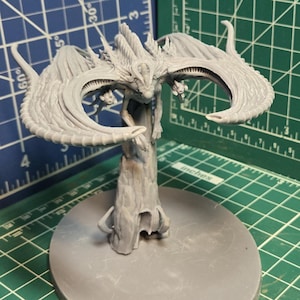 May include: A grey, unpainted resin miniature of a winged creature perched atop a vertical, textured pillar. The figure features large, curved wings extending outwards and is mounted on a smooth, oval-shaped base, captured against a grid mat.