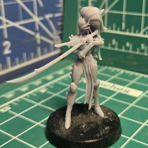 May include: A gray unpainted miniature figurine of a humanoid character standing on a circular black base. The figure features long hair, armor plating, and holds a long, slender sword extended forward in a combat stance.